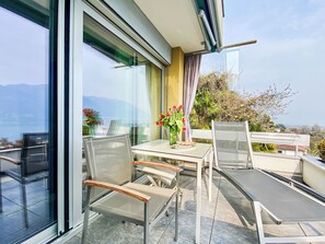 A modern outdoor seating area with glass walls, a table with chairs, and a vase of flowers. - Lakeview App 501 by Interhome (Ascona)