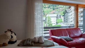 A hotel room with a bed, a red leather sofa, and a view of a mountainous landscape through large windows.