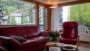 A modern living room with a red leather sofa, a red armchair, and a small table with a vase of flowers. Large windows offer a view of a mountainous village.
