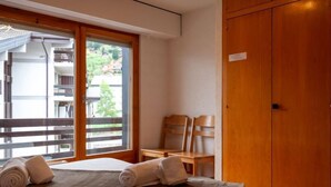 A hotel room with a bed, a wooden wardrobe, a window with a view of a balcony, and two chairs.