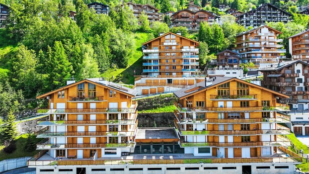 Un hotel de varios pisos con balcones, con un fondo de exuberante vegetación y otros edificios.