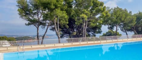 En klar, blå swimmingpool med træterrasse og metalgelænder, omgivet af høje træer og udsigt til havet.