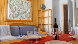 Une pièce chaleureuse avec un mur en bois, une grande carte topographique, un canapé, une table à manger dressée avec des assiettes et des verres, et une bouteille de vin.