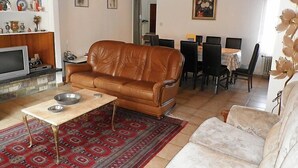 Ein Wohnzimmer mit braunem Ledersofa, einem weißen Sofa, einem Essbereich mit Tisch und Stühlen sowie einem Fernseher auf einem Lowboard.