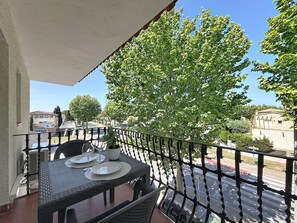 Un balcon avec une table dressée pour deux personnes, un pot de fleurs et une vue sur une rue bordée d’immeubles et d’arbres.