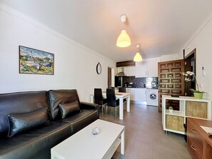 A modern living room with a black leather sofa, a white coffee table, and a dining area with a table and chairs. The kitchen is visible with white cabinets and a stove.