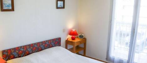 Un dormitorio con una cama, una mesita de noche con una lámpara y un despertador, una ventana con vistas al exterior, y dos cuadros en la pared.