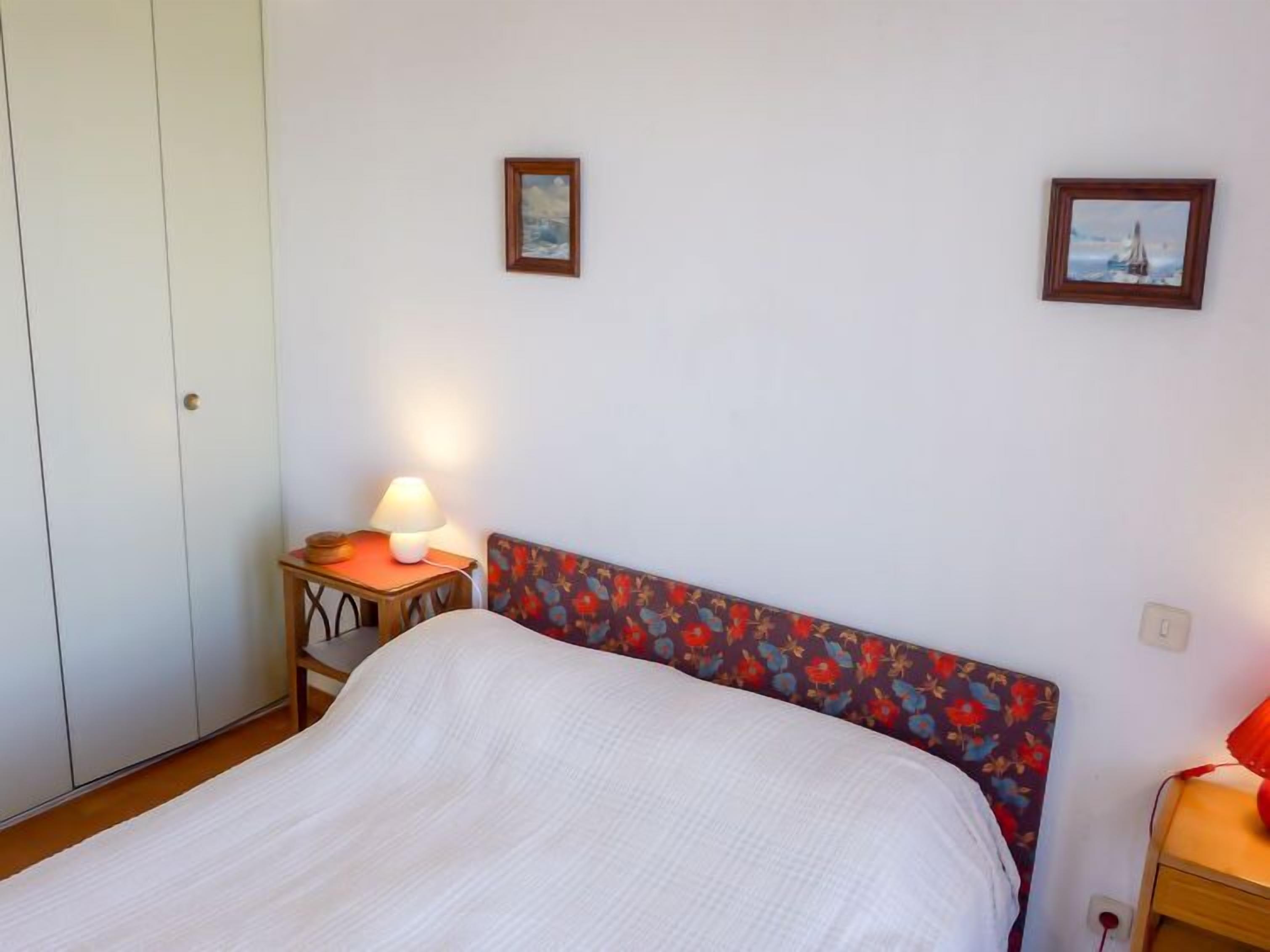 A bedroom with a bed, a nightstand with a lamp, a wardrobe, and two framed pictures on the wall.