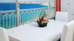 Un balcon avec une table et des chaises blanches, un panier en osier rempli de fleurs et une vue sur le paysage urbain.