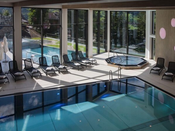A modern indoor pool area with clear glass walls, lounge chairs, and a hot tub. - Goldried Park by Interhome (Matrei in Osttirol)