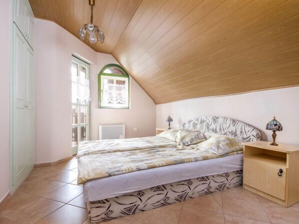 A bedroom with a sloped ceiling, a bed with patterned bedding, a bedside table with lamps, and a window with a green frame. - Vacation home Green by Interhome (Balatonmariafurdo)