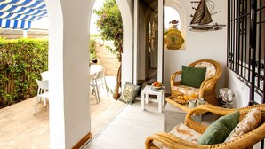 A room with a view of the garden, a wicker sofa, a dining table with chairs, and a decorative sailboat wall art piece. - Pueblo Andaluz by Interhome (Dénia)