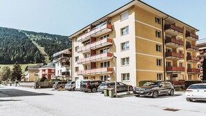 Um edifício de apartamentos com várias andares, varandas, carros estacionados e montanhas ao fundo.