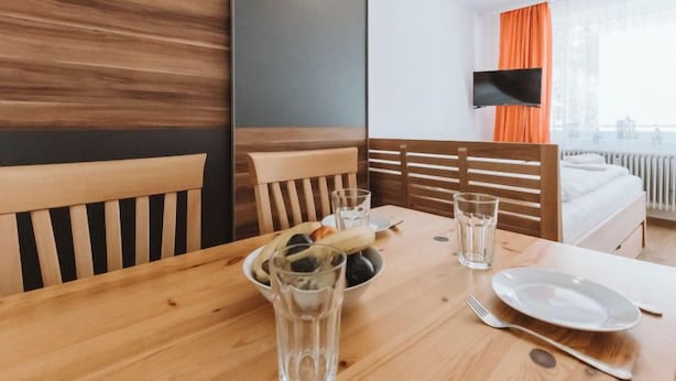 Quarto de hotel com mesa de jantar de madeira posta para dois, cama com roupa de cama branca e televisão fixada na parede.