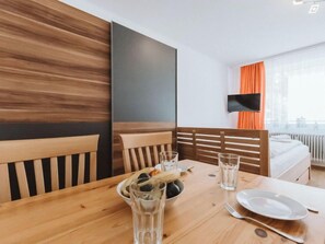 Habitación de hotel con una mesa de comedor de madera dispuesta para dos personas, una cama con ropa blanca y un televisor montado en la pared.
