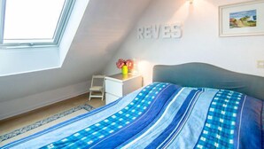 Une chambre avec un plafond en pente, un lit recouvert d’une couvre-lit à rayures bleues et blanches, une petite table de chevet blanche avec une lampe, un tableau encadré accroché au mur et une fenêtre laissant entrer la lumière naturelle.