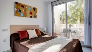 Une chambre d’hôtel avec un grand lit, une table de chevet, une lampe et une porte-fenêtre coulissante donnant sur un balcon aménagé avec du mobilier d’extérieur.
