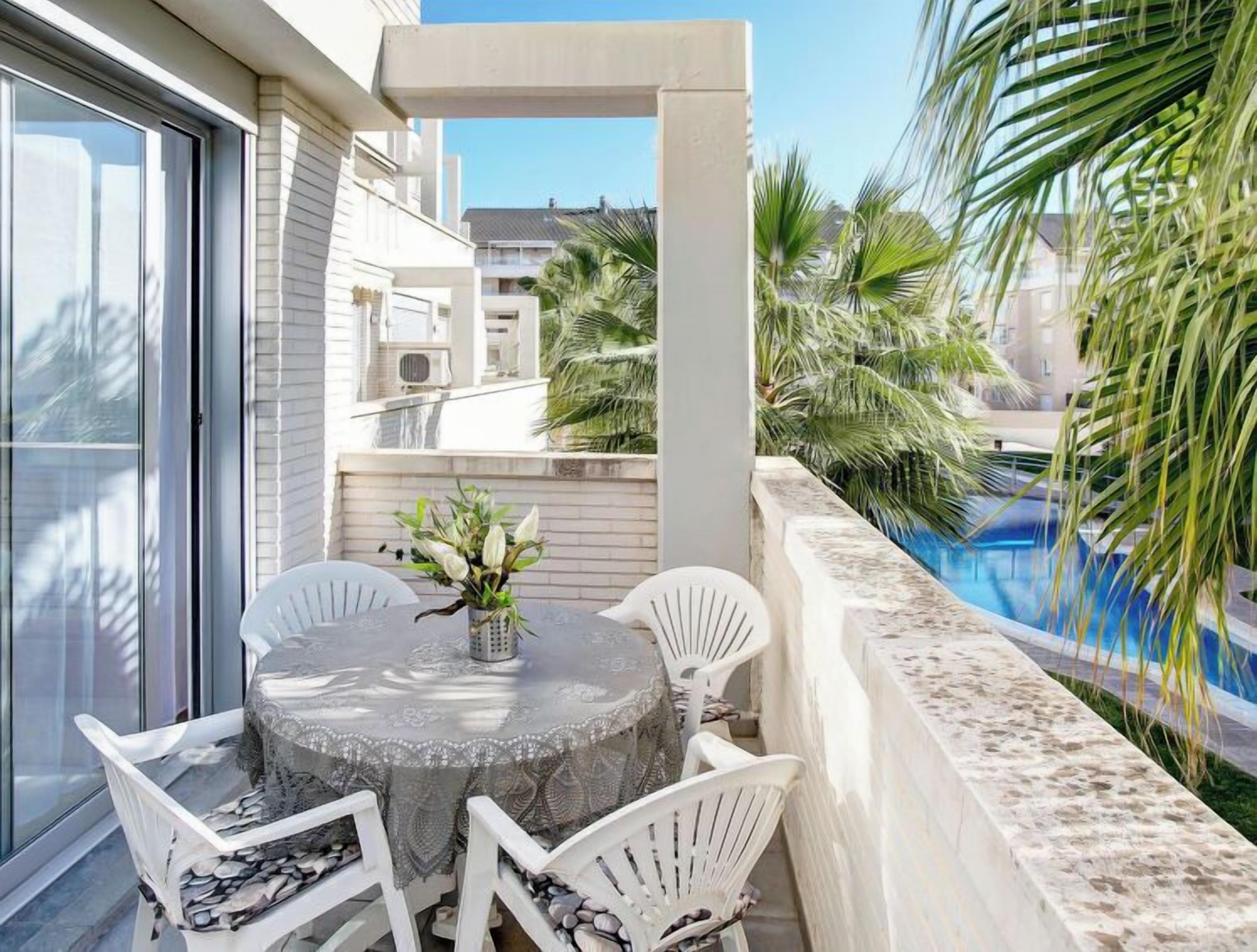A balcony with a round table and chairs, a small vase with flowers, and a pool in the background.
