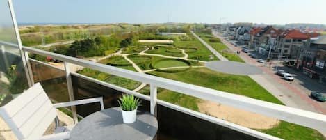 A balcony with a table and chairs, overlooking a landscaped garden and a residential area with houses and a road.