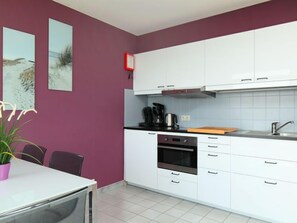 A modern kitchen with white cabinets, a black oven, and a coffee maker. There is a dining table with chairs and a potted plant.