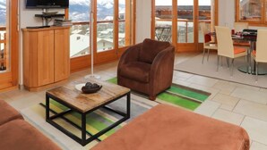 A modern living room with a sofa, a brown armchair, a coffee table, and a TV. Large windows offer a view of a snowy mountain range. - Domaine des Reines B4 by Interhome (Nendaz)