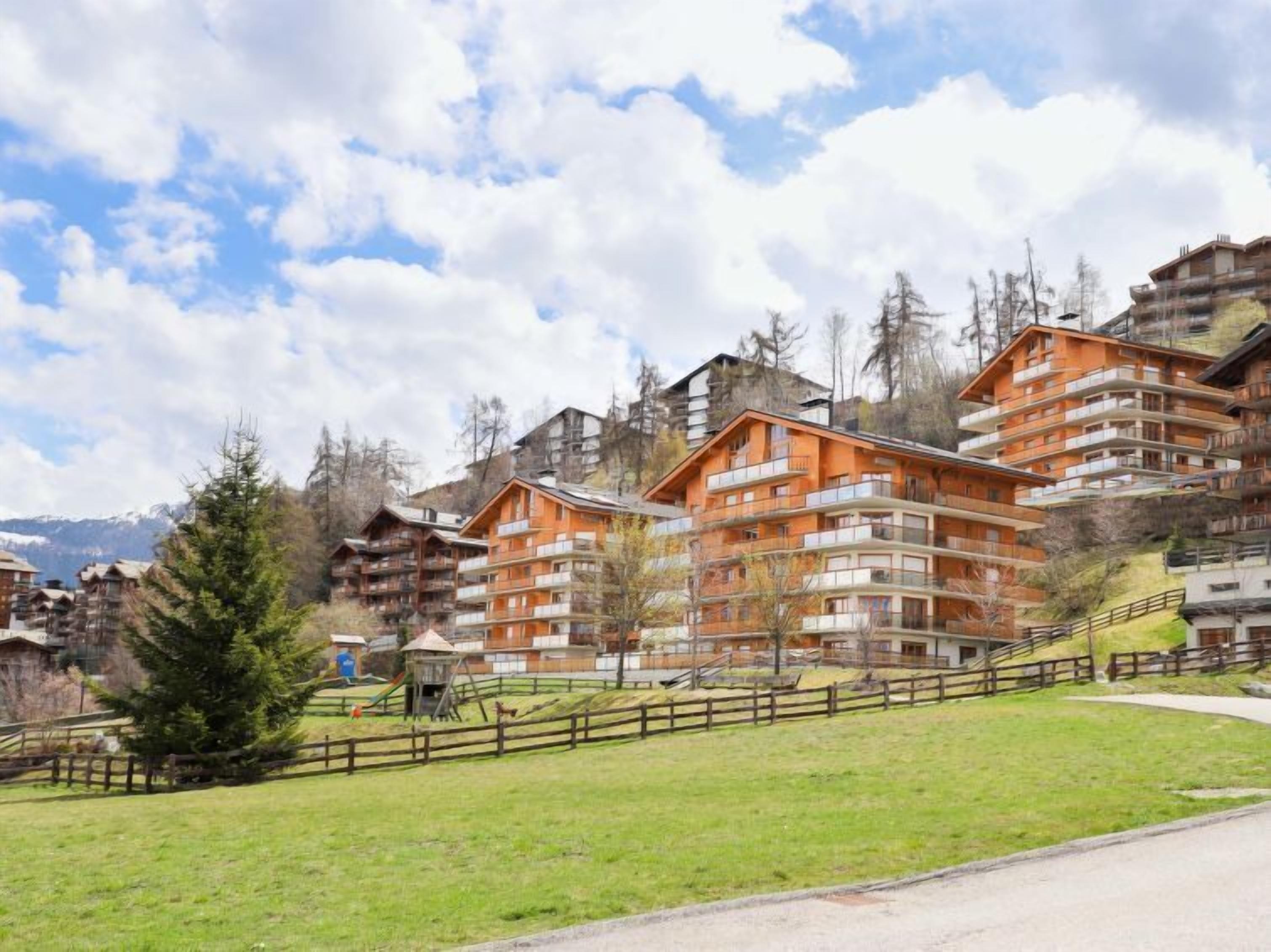 Un ensemble d’immeubles à plusieurs étages, dotés de balcons, se dressant sur un fond de montagnes et sous un ciel partiellement nuageux.