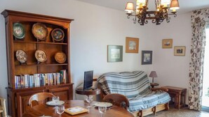 Un comedor con una mesa de madera dispuesta para cuatro personas, rodeada de sillas. A la izquierda, una estantería llena de libros y platos decorativos. En el fondo, un sofá con una manta a rayas, y un candelabro colgando encima.