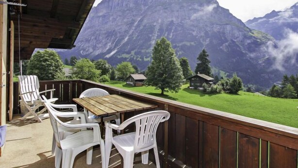 Eine Holzterrasse mit Tisch und weißen Plastikstühlen, mit Blick auf eine Berglandschaft mit Häusern und Bäumen.