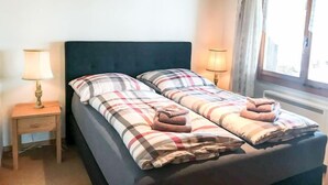 Ein Bett mit kariertem Überwurf und zwei Handtüchern, eine Nachttischlampe und ein Fenster mit Vorhängen.