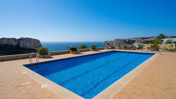 A rectangular pool with a clear blue sky and ocean view, surrounded by a tiled deck and potted plants. - Pueblo de la Luz by Interhome (Benitachell)