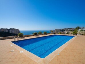 A rectangular pool with a clear blue sky and ocean view, surrounded by a tiled deck and potted plants. - Pueblo de la Luz by Interhome (Benitachell)
