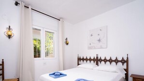 A bedroom with a wooden bed, white curtains, a window with a view of greenery, and a wall-mounted light fixture. - Pueblo de la Luz by Interhome (Benitachell)