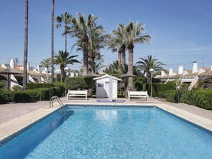 A swimming pool with a small structure in the center, surrounded by palm trees and white buildings with thatched roofs.
