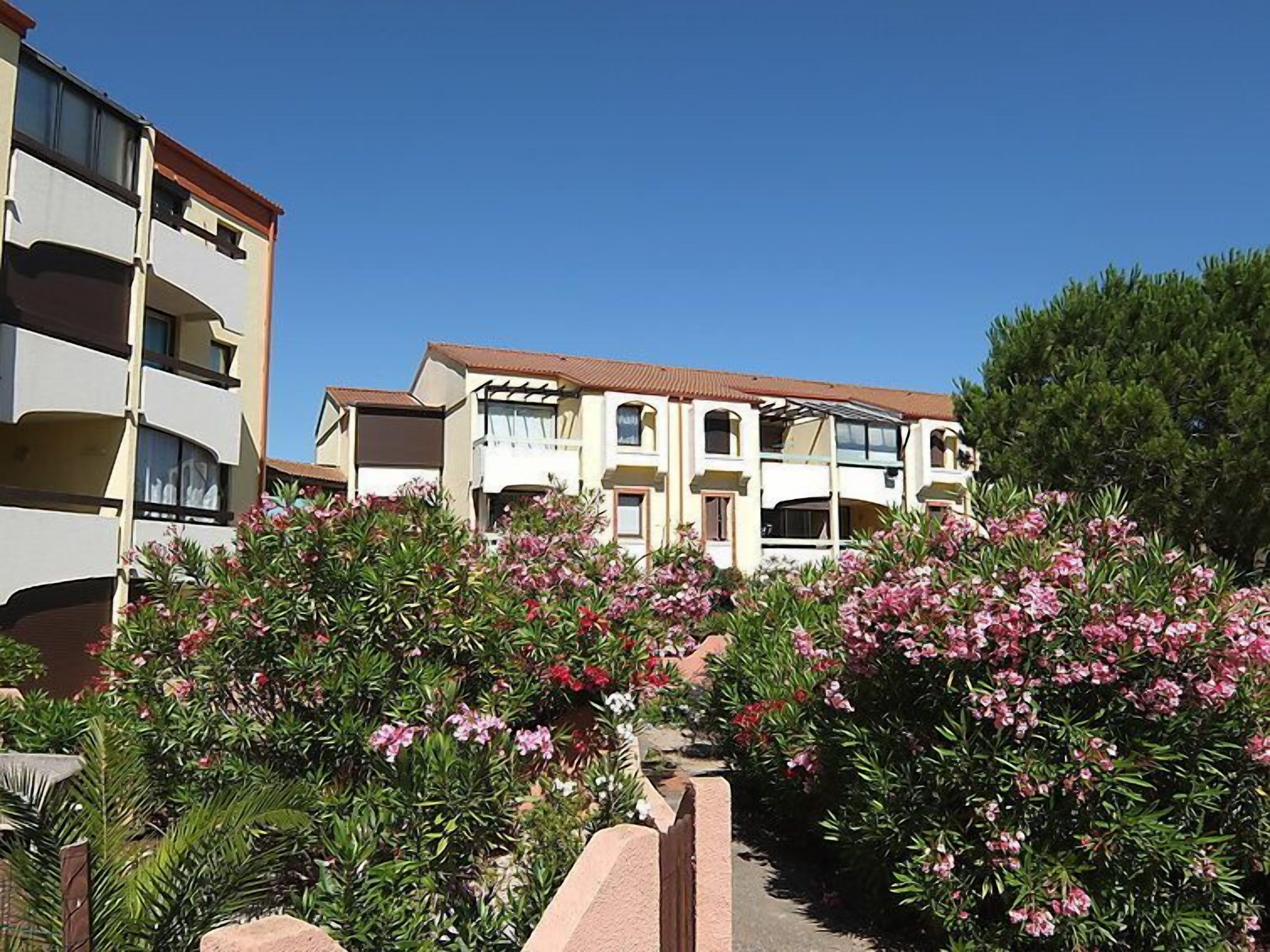 A multi-story residential building with balconies, surrounded by lush greenery and flowering plants.