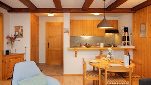 A cozy room with a blue armchair, a wooden dining table with chairs, a kitchen area with wooden cabinets, and a hanging light fixture. - Parc C013 by Interhome (Champex)