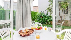 A breakfast table set with a basket of pastries, a bowl of fruit, and glasses of orange juice, surrounded by white chairs and a glass window overlooking a garden. - Club Sevilla I by Interhome (Oliva)