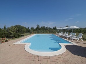 A rectangular swimming pool with a curved edge, surrounded by a brick patio. There are white lounge chairs and umbrellas along the poolside.