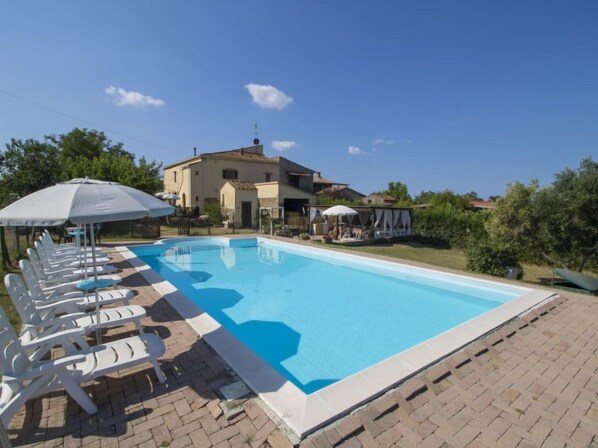 A swimming pool with lounge chairs and umbrellas, surrounded by a brick patio and a building with a tiled roof.