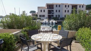 An outdoor seating area with a table and chairs, surrounded by greenery, overlooking a marina with boats and a building in the background. - Marina 2 by Interhome (Saint Cyprien)