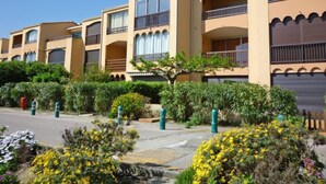 A row of beige multi-story buildings with balconies, surrounded by greenery and yellow flowers. - Marina 2 by Interhome (Saint Cyprien)