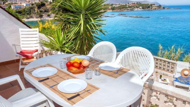 A round table with white chairs and a fruit bowl, overlooking a coastal view with buildings and mountains. - Nicolas by Interhome (Llançà)