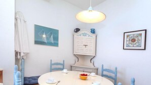 A dining area with a round table, blue chairs, and a white cabinet with a clock above it. - Nicolas by Interhome (Llançà)