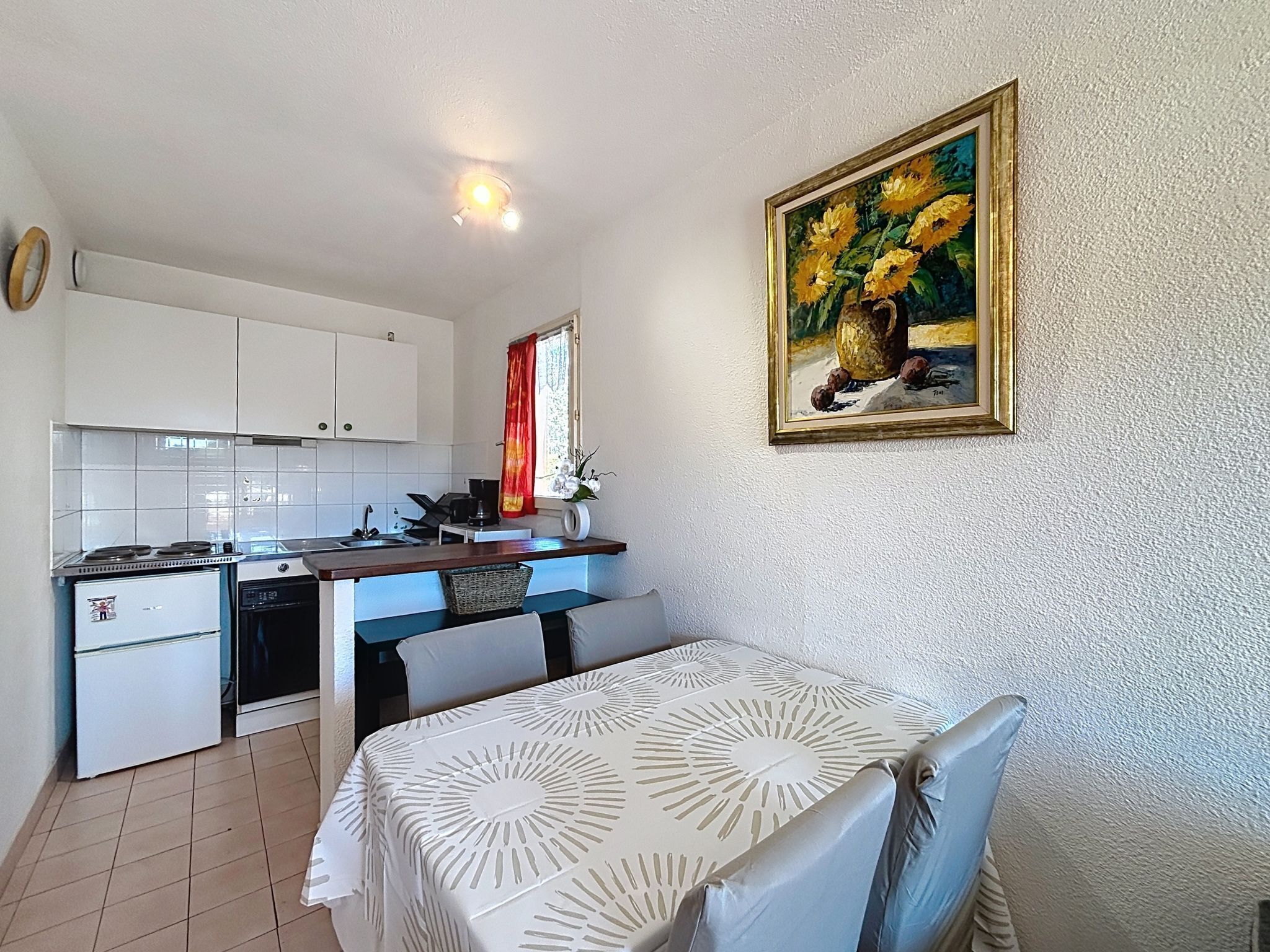 Une cuisine de taille réduite avec des meubles blancs, une table à manger recouverte d’une nappe blanche et un tableau représentant des fleurs accroché au mur.