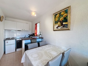 A compact kitchen with white cabinets, a dining table with a white tablecloth, and a painting of flowers on the wall.