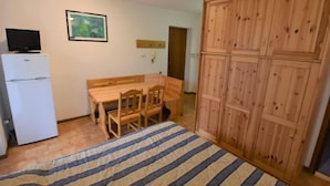 Una habitación con cama, una mesa de comedor de madera con sillas, un refrigerador blanco y un armario de madera.