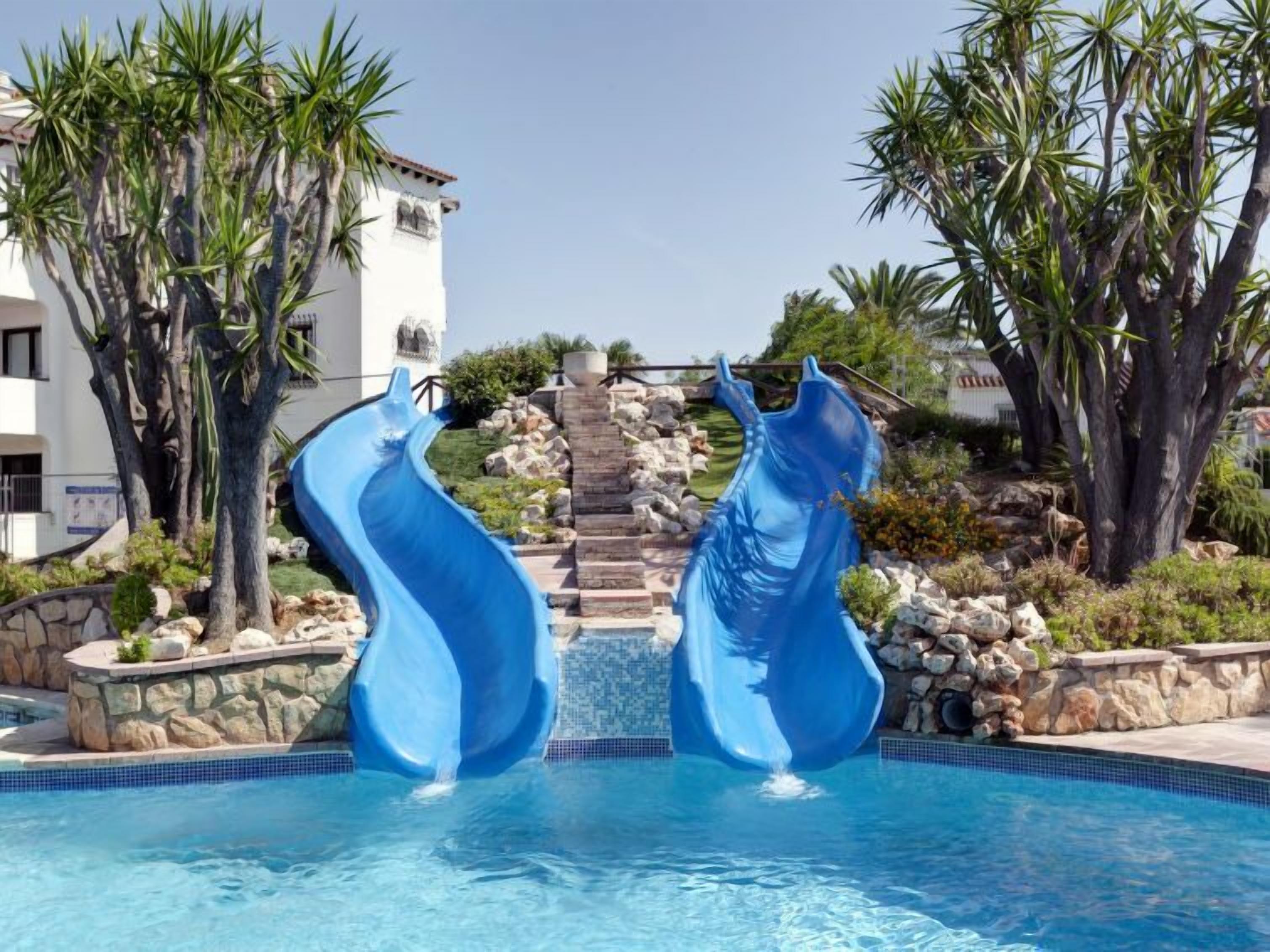 Une piscine dotée de deux toboggans bleus, entourée de palmiers et accessible par un escalier en pierre menant à un bâtiment.