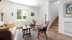 Ein Wohnzimmer mit einer Couch, zwei Korbstühlen, einem Couchtisch und einem Fenster mit Blick ins Grüne.