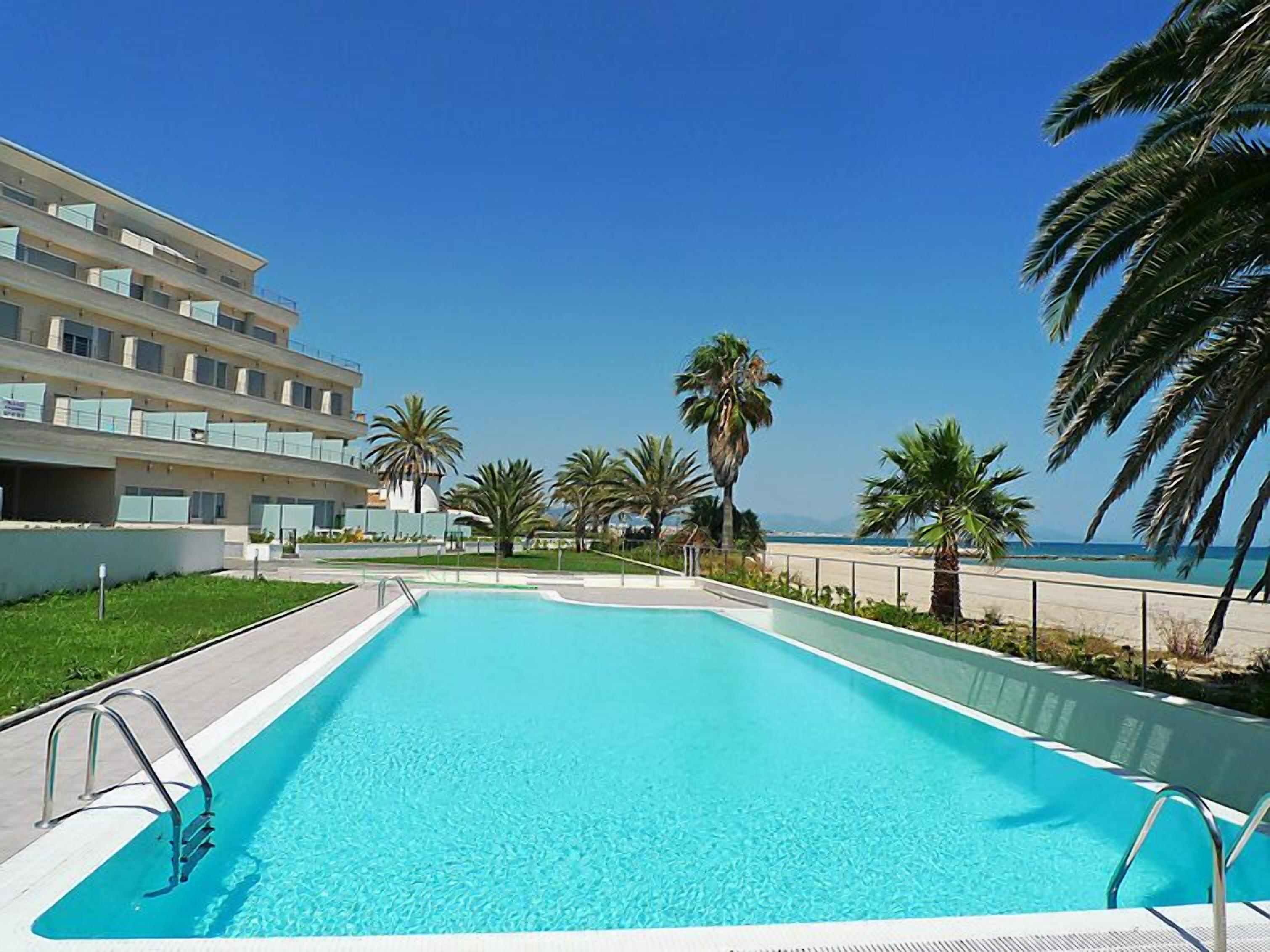 A swimming pool with a clear blue water, surrounded by palm trees and a building with balconies.