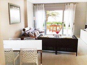 A modern living room with a sofa, a dining table, and chairs. There is a large window with curtains, and a view of the outdoors.
