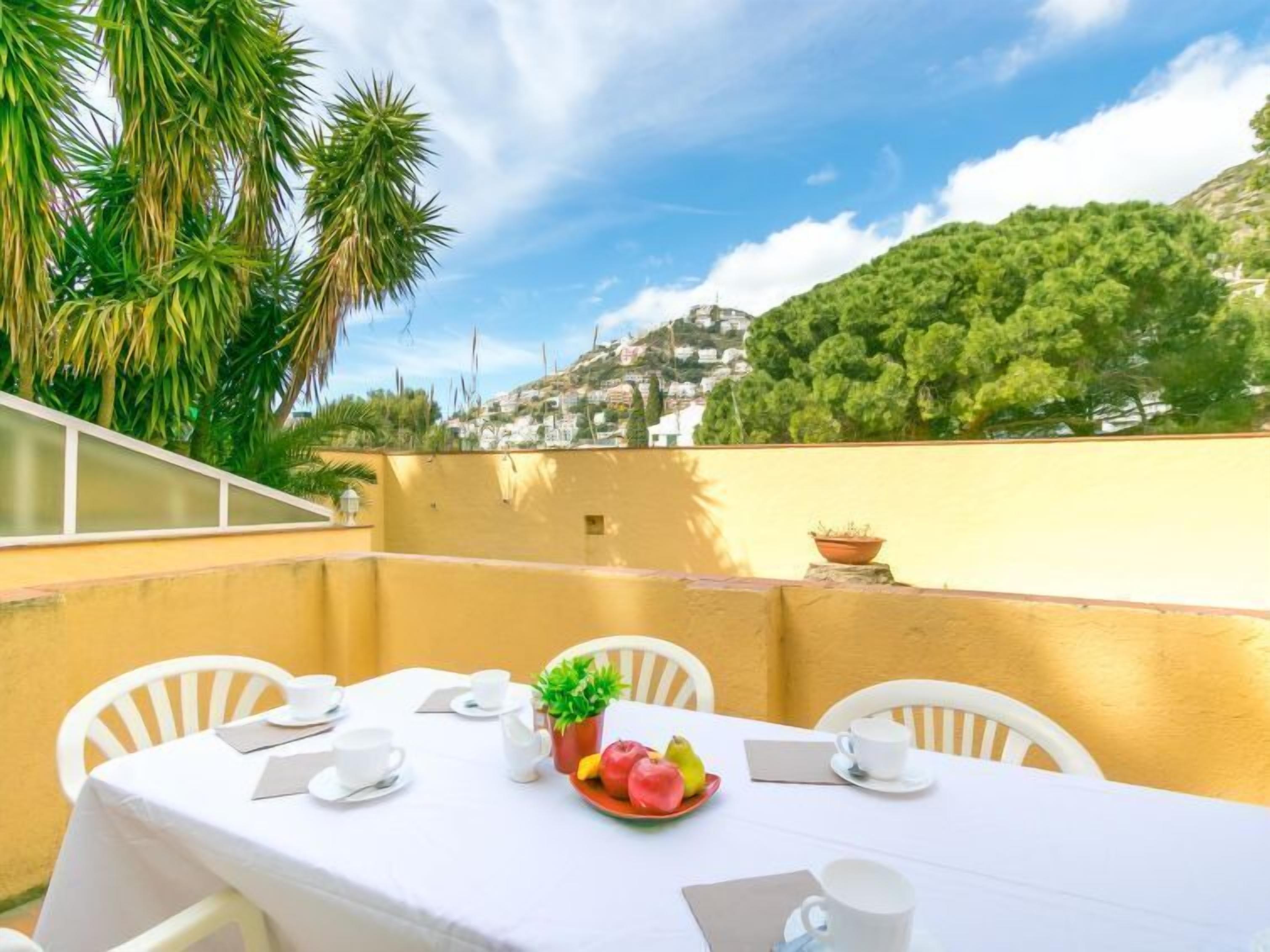 A table set for breakfast with white chairs, a yellow wall, and a view of a hillside with buildings.
